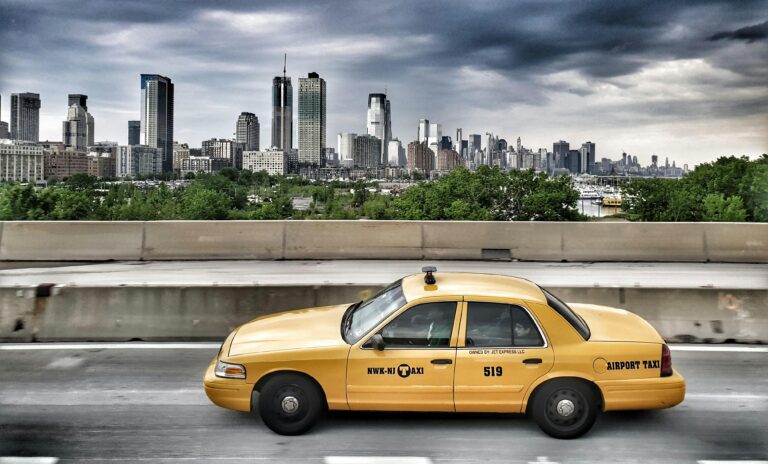 Iconic yellow taxi driving with New York City skyline in the background under a dramatic sky.